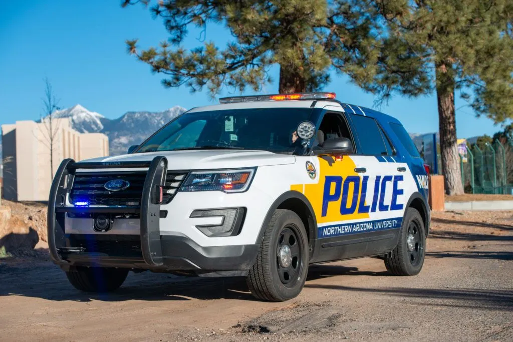 NAU Police Department vehicle parked under pine trees with the San Francisco Peaks behind it